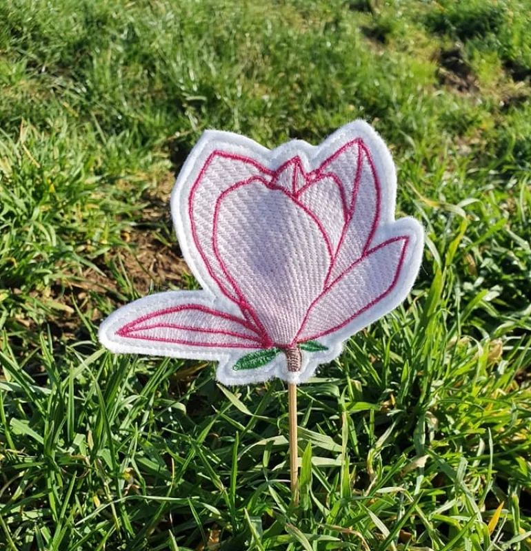 Freebie Stickdatei ITH Stabdatei Magnolie, Foto einer Ausfertigung, die mit dem Stab in einer grünen Wiese steckt. Motiv: Magnolienblüte mit Stielansatz und zwei grünen Blättchen. Untergrundstoff ist heller Filz in Blütenumriss in roten Konturen mit zarte