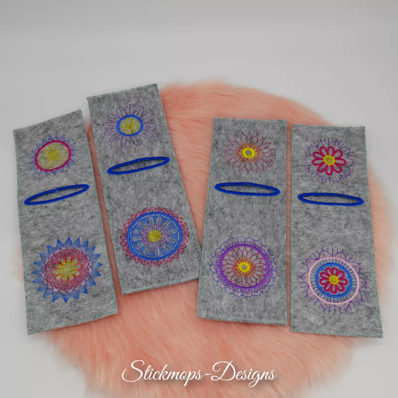 Stickdatei Set Mandala-Bestecktaschen + Einzelmotive, Foto mit 4 mittelgrauen Filztaschen, auf orangefarbenem Fell liegend. Jede mit blau umstickter Öffnung im oberen Drittel und jeweils unterschiedlichen Bestickungen oberhalb und unterhalb davon.
Je nach