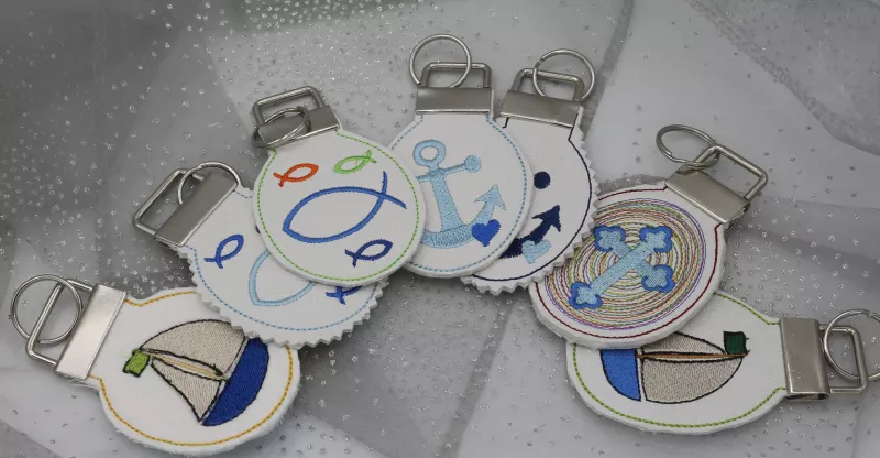 Stickdatei Set 4 Schlüsselanhänger mit christlichen Symbolen. Foto zeigt 7 ovale Anhänger mit je geklammerter Öse, durch die ein Schlüsselring gezogen ist. Mit den Motiven: Segelboot, Anker, Kreuz, Fischsymbole.