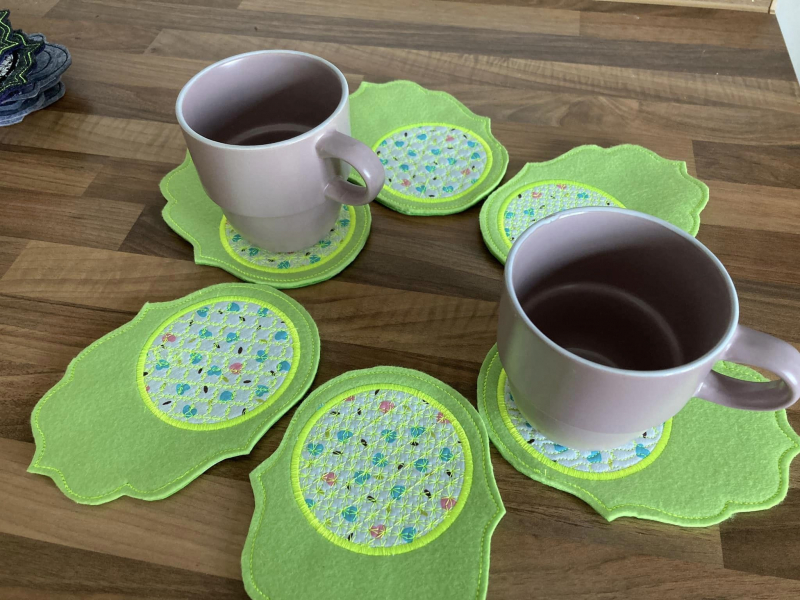 Stickdatei Set ITH Mug Rug 7, sechs mal ausgestickt und kreisförmig drapiert auf einem Tisch, wobei auf zwei von ihnen je eine Tasse steht. Die Mug Rugs zeigen sich auf lindgrünem Filzuntergrund, welcher zu einer Seite länglich in einem welligen Schnörkel