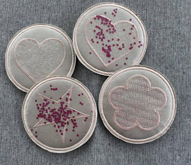 Stickdatei Set ITH Mug Rugs und Anhänger, Foto zeigt 4 kreisrunde Mug Rugs silbrig-braunen Untergrunds. Alle mit dezentem Symbol (Blüte, Herz, Stern) und ebensolcher Innenbestickung. 2 davon mit eingearbeiteter perlchenartiger Bestreuung in rot. Stickdatei Set ITH Mug Rugs und Anhänger, Foto zeigt 4 kreisrunde Mug Rugs silbrig-braunen Untergrunds. Alle mit dezentem Symbol (Blüte, Herz, Stern) und ebensolcher Innenbestickung. 2 davon mit eingearbeiteter perlchenartiger Bestreuung in rot.