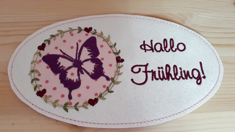 Stickdatei Set Hallo Frühling (div. Rahmengrößen), Foto zeigt ovalen hellen Mug Rug. Linksseitig lila Schmetterling auf rosa-pink gemustertem Stoff, umgeben von Kranz aus Herzen und Blättern. Rechts Schriftzug "Hallo Frühling!".