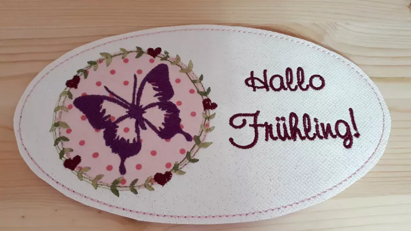 Stickdatei Set Hallo Frühling (div. Rahmengrößen), Foto zeigt ovalen hellen Mug Rug. Linksseitig lila Schmetterling auf rosa-pink gemustertem Stoff, umgeben von Kranz aus Herzen und Blättern. Rechts Schriftzug "Hallo Frühling!".