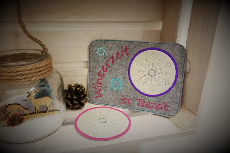 Freebie Stickdatei ITH Mug Rug Winterzeit (13x18 und 10x10 Rahmen), Foto mit komplettem querformatigem Tassenteppich nebst einzelnem rundem Abstellplatz, umgeben von beigefügten Deko-Elementen. Teppich grau, Abstellplatz beige mit lila Rand und hellem Eis
