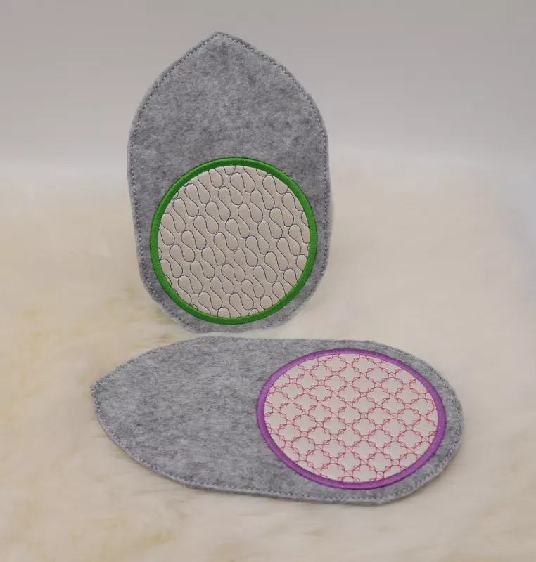 Stickdatei Set ITH Mug Rug 5, Foto mit einem stehenden und einem liegenden Untersetzer. Diese sind als kreisrundes dick umrandetes Zentrum auf einem hellgrauen Filzuntergrund gestickt, dessen Oval zur einen Seite  spitz ausgezogen ist. Das Zentrum ist auf