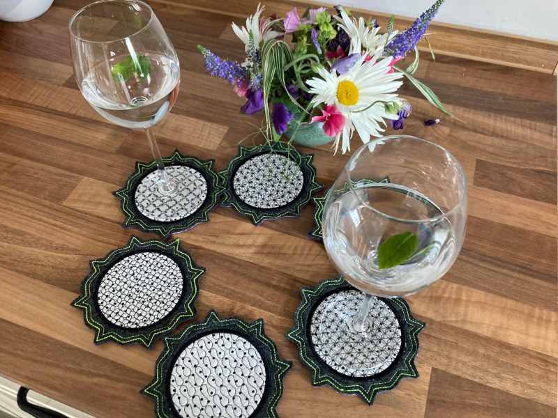 Stickdatei Set ITH Mug Rug 6, Tischarrangement mit kleinem buntem Blumenstrauß und sechs kreisförmig ausgelegten Mug Rugs, wobei zwei von ihnen je ein Weinglas tragen. Alle sechs haben einen strahlenförmig und doppelt am Rand abgesteppten dunkelgrauen Fi Stickdatei Set ITH Mug Rug 6, Tischarrangement mit kleinem buntem Blumenstrauß und sechs kreisförmig ausgelegten Mug Rugs, wobei zwei von ihnen je ein Weinglas tragen. Alle sechs haben einen strahlenförmig und doppelt am Rand abgesteppten dunkelgrauen Fi
