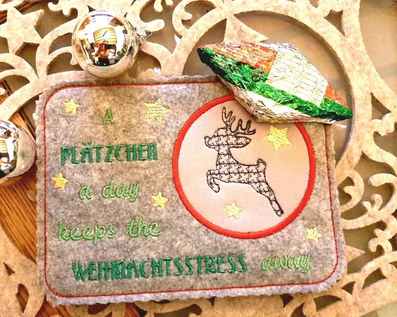 Stickdatei Set ITH Reh-Deko Sparset 4 in 1, Foto mit querformatigem Tassenteppich aus grauem Filz, weihnachtliche Dekoration. Teppich hat kreisrunden Abstellplatz mit Bestickung Reh/Sterne. Außen, umgeben von Sternen, grüne Aufschrift: "A PLÄTZCHEN a day