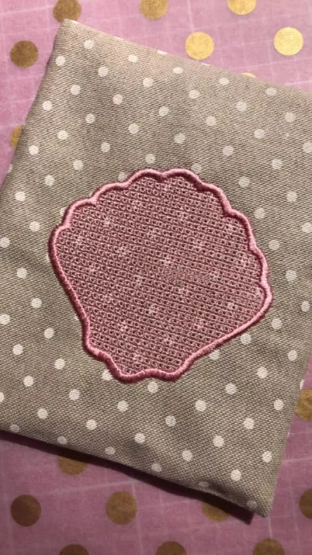 Stickdatei Set Maritime Handtuch Motive Embossed 10x10, Foto zeigt graubraunen Stoff, weiß gepunktet, mit Stickemblem. Motiv: Muschelform, dick umrandet, Füllfläche filigran gemustert, Farbe uni altrosa.