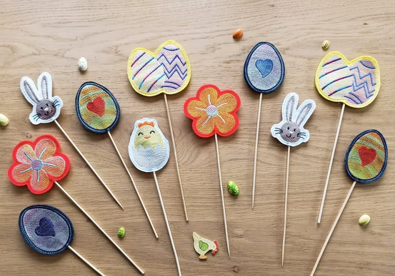 Stickdatei Set Oster-Stabdesigns, Foto mit 11 Designs, auf Holzuntergrund liegend, dazwischen kleine Dekoelemente als Beigaben. 4 mal Ei mit Herz darin; 2 mal Hasenkopf; 1 mal Küken in Eierschale; 2 mal Blüten;  2 mal Doppel-Osterei.