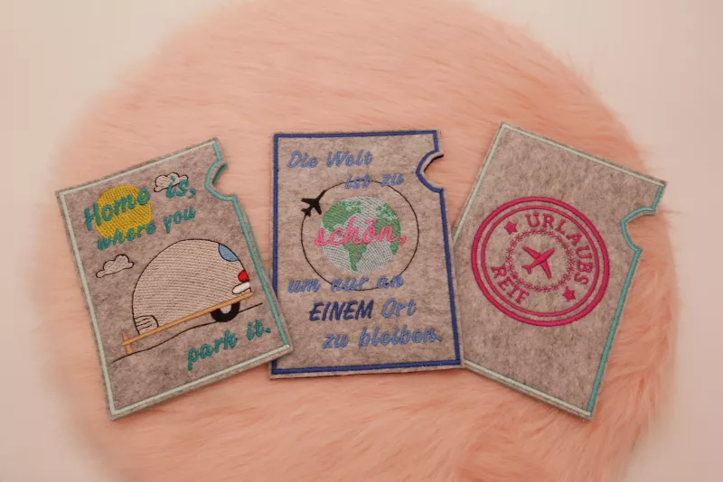 Stickdatei Set ITH Reisepasshüllen (13x18) + Einzelmotive, Foto mit 3 Hochkant-Hüllen aus hellgrauem Filz. Diverse bunte Bestickungen mit urlaubsbezogenem Motiv und Schriftzug ("Home is where you park it", "Die Welt ist zu schön, um nur an einem Ort zu bl