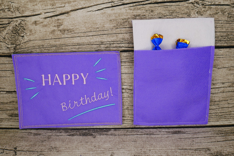 Stickdatei Set ITH Geburtstagskarten (10x10 u. 13x18), Foto mit 2 lila Karten. Karte im Hochkantformat zeigt ihre Rückseite, die eine Einstecktasche aufweist, aus der 2 Bonbonpapierspitzen ragen.
Karte im Querformat zeigt Bestickung: "HAPPY Birthday!"/Str