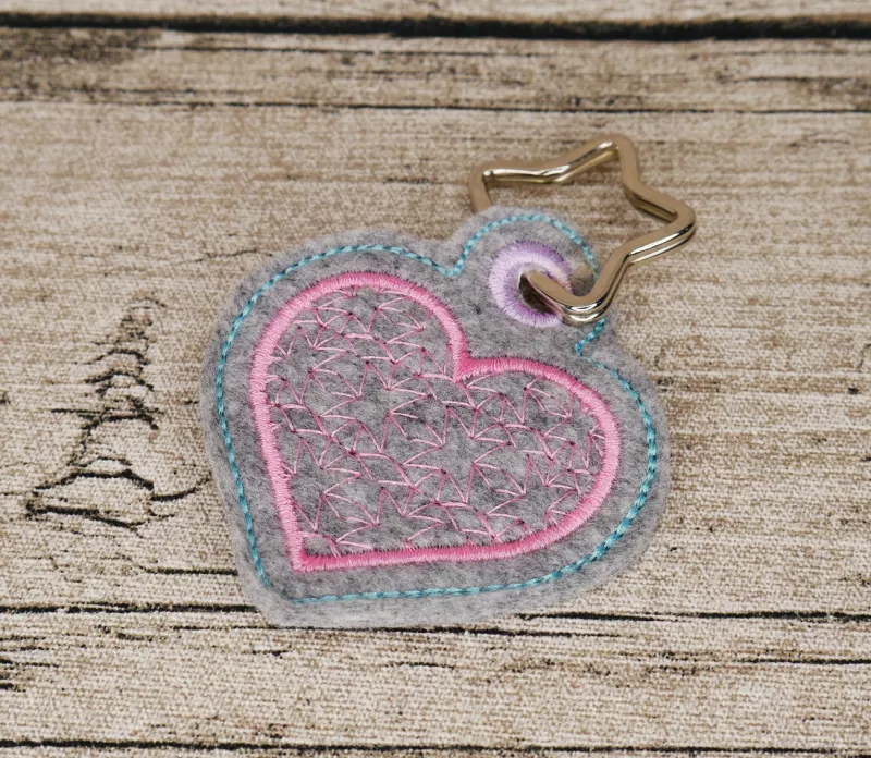 Stickdatei Set ITH Mug Rugs und Anhänger, Foto zeigt Anhänger in Herzform, oben geöst mit durchgezogenem sternförmigem Schlüsselring. Grauer Filz, rosa Herz mit ebensolcher Umrandung und filigraner Innenbestickung gleicher Farbe.