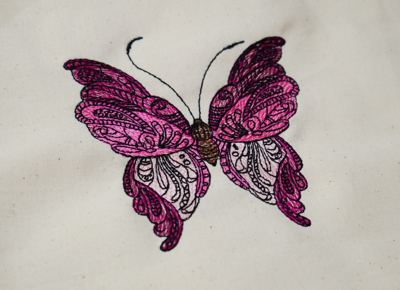 Stickdatei Schmetterling Vollstickmotiv, Foto mit Stickerei auf hellem Stoff. Schmetterling in pink-rosa-roten Farben mit schwarzem filigranem Muster darauf.