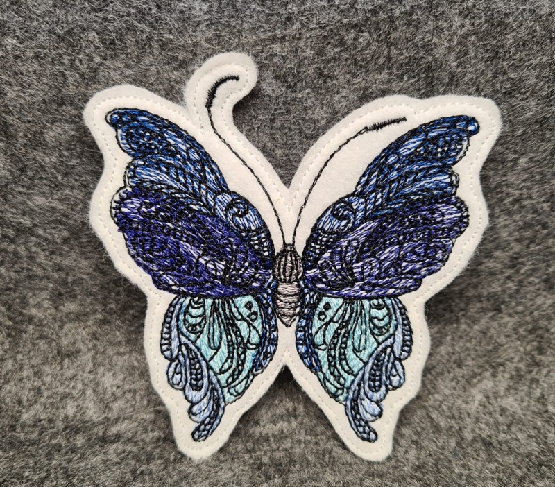 Stickdatei Schmetterling Vollstickmotiv, Foto mit Ausfertigung auf dunkelgrauem Untergrund. Motiv: Schmetterling in lila-blau-türkis-schwarz.