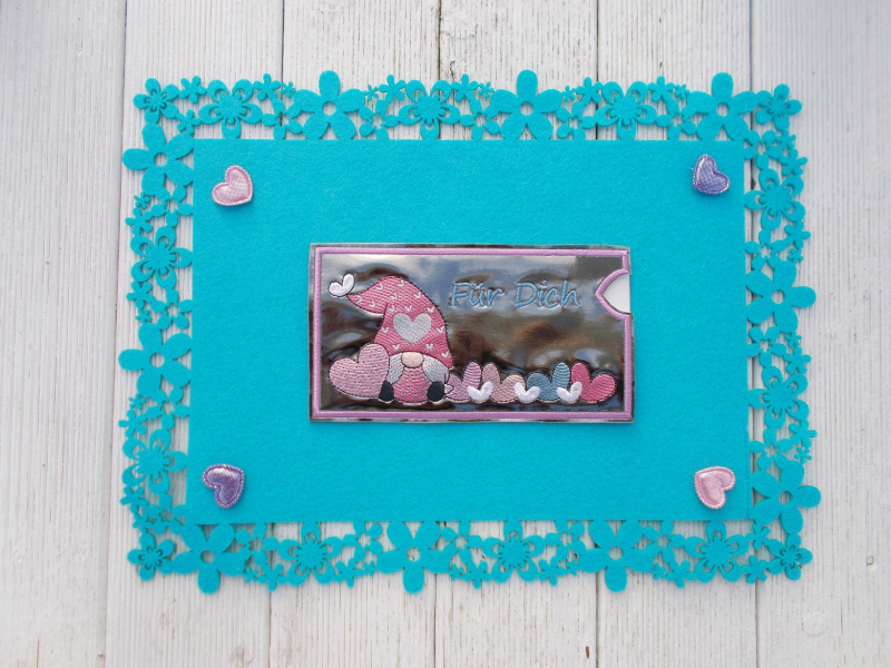 Stickdatei Set Lovegnomes ITH Geschenk- und Schokoladenhüllen, Foto mit 1 Ausfertigung, präsentiert auf türkisem Deckchen, das auf Holzuntergrund liegt. Dekoherzchen als Beilagen an den Deckenecken. Querformatige glänzende Kunstleder-Hülle mit Bestickung: Stickdatei Set Lovegnomes ITH Geschenk- und Schokoladenhüllen, Foto mit 1 Ausfertigung, präsentiert auf türkisem Deckchen, das auf Holzuntergrund liegt. Dekoherzchen als Beilagen an den Deckenecken. Querformatige glänzende Kunstleder-Hülle mit Bestickung: