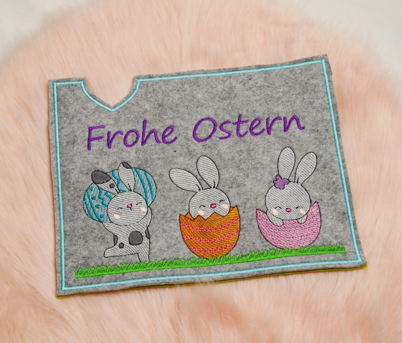 Stickdatei Set Osterhüllen ITH Geschenk- und Schokoladenhüllen + Einzelmotive. Foto, das eine Ausfertigung auf apricotfarbenem Fell liegend zeigt. Querformat-Hülle aus grauem Filz, Schriftzug "Frohe Ostern", darunter 3 verschiedene Osterhasenmotive mit Ei