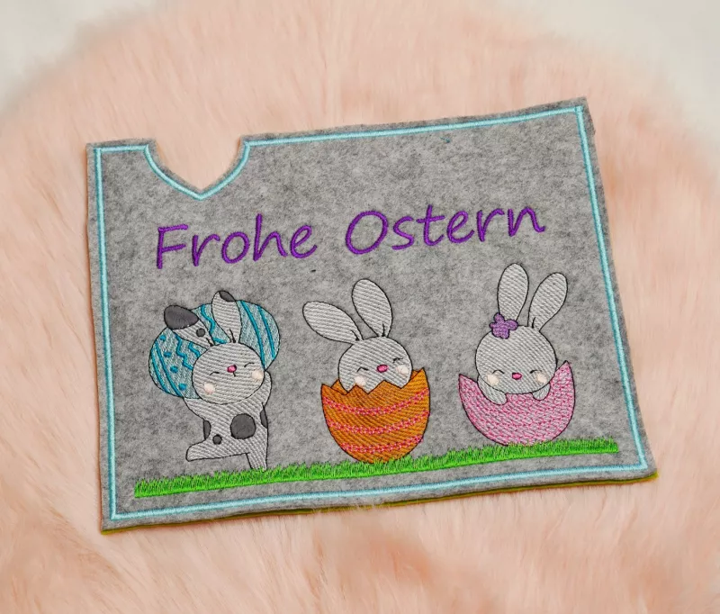 Stickdatei Set Osterhüllen ITH Geschenk- und Schokoladenhüllen + Einzelmotive. Foto, das eine Ausfertigung auf apricotfarbenem Fell liegend zeigt. Querformat-Hülle aus grauem Filz, Schriftzug "Frohe Ostern", darunter 3 verschiedene Osterhasenmotive mit Ei