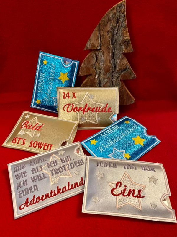 Stickdatei Set ITH Hüllen für Adventskalender to go, Foto mit 6 querformatigen Hüllen in diversen glänzenden/glitzerigen Farben auf rotem Untergrund, Weihnachtsdeko aus Holz im Hintergrund. Bestickung: Rentier, bzw. Stern, bzw. Weihnachtsbaum. Schriftzüge