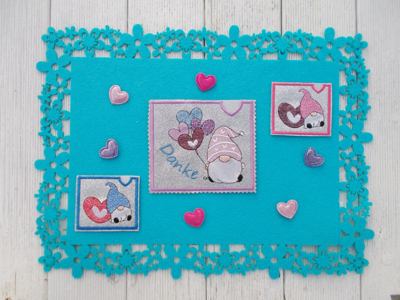 Stickdatei Set Lovegnomes ITH Geschenk- und Schokoladenhüllen, Foto mit 3 Ausfertigungen, präsentiert auf türkisem Deckchen, das auf Holzuntergrund liegt. Dekoherzchen als Beilagen dazwischen. 3 Querformatige Hüllen aus grauem Filz mit Bestickung: 2 mal G Stickdatei Set Lovegnomes ITH Geschenk- und Schokoladenhüllen, Foto mit 3 Ausfertigungen, präsentiert auf türkisem Deckchen, das auf Holzuntergrund liegt. Dekoherzchen als Beilagen dazwischen. 3 Querformatige Hüllen aus grauem Filz mit Bestickung: 2 mal G