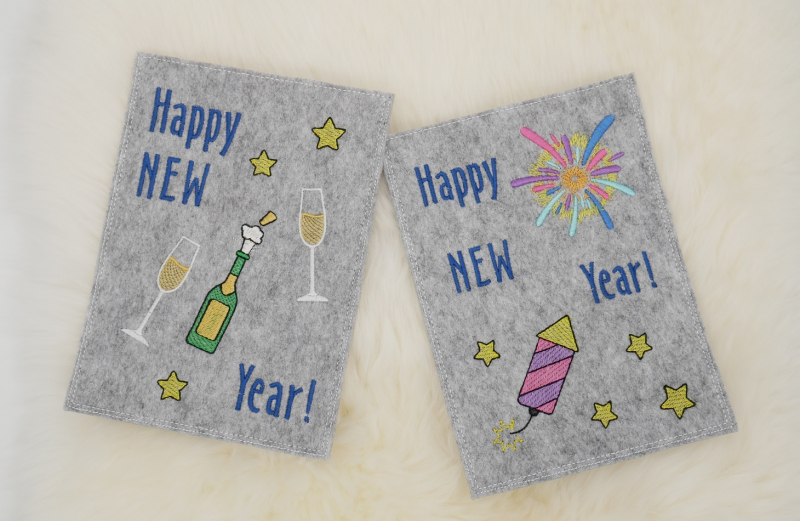 Stickdatei Set Gnom Silvesterparty mit ITH Kartenhüllen & Einzelmotive, Foto zeigt 2 Hochkant-Karten aus hellgrauem Filz. Eine mit Sternen, Sekt und Gläsern, Schriftzug "Happy New Year!". Eine mit Raketen, Sternen, Feuerwerksstrahlen, Schriftzug "Happy N