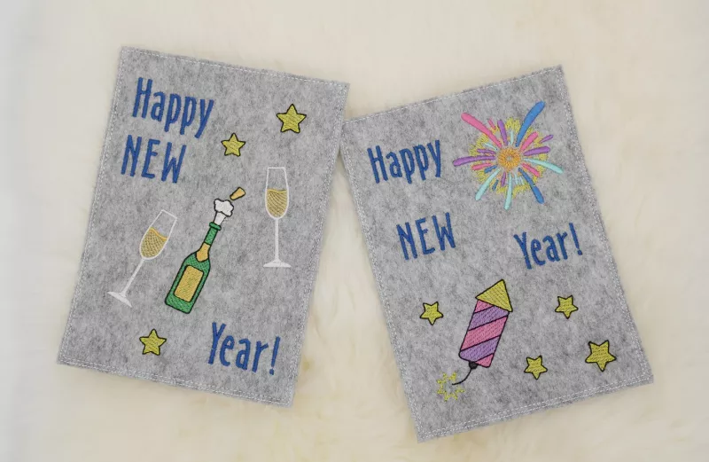 Stickdatei Set Gnom Silvesterparty mit ITH Kartenhüllen & Einzelmotive, Foto zeigt 2 Hochkant-Karten aus hellgrauem Filz. Eine mit Sternen, Sekt und Gläsern, Schriftzug "Happy New Year!". Eine mit Raketen, Sternen,  Feuerwerksstrahlen, Schriftzug "Happy N