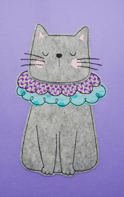 Stickdatei Set Little Sweet Cat Doodle Applikation inkl. ITH Anhänger, Foto zeigt Applikation auf lila Stoff. Sitzende graue Katze mit Halskrause in lila- und türkis-blau-gemustert.