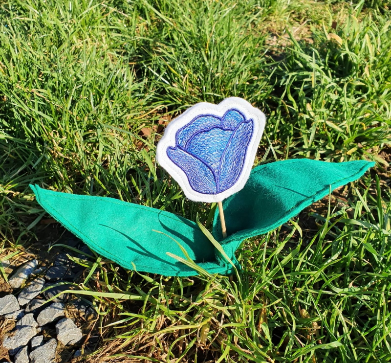 Stickdatei ITH Tulpe (10x10 und größer), Foto zeigt in Rasen gesteckten Designstab. Tulpenkopf in Blautönen auf weißem Untergrund. Am Stab befestigt 2 grüne Blätter, aus Filz genäht.