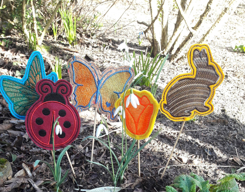 Stickdatei Set ITH Frühlings-Stabdesigns, Foto zeigt 5 Designstäbe, im Boden eines Außenbereichs steckend. Unterschiedliche bunte Bestickungen, Motive: Marienkäfer, Hase, Schmetterlinge, Tulpenblüte.