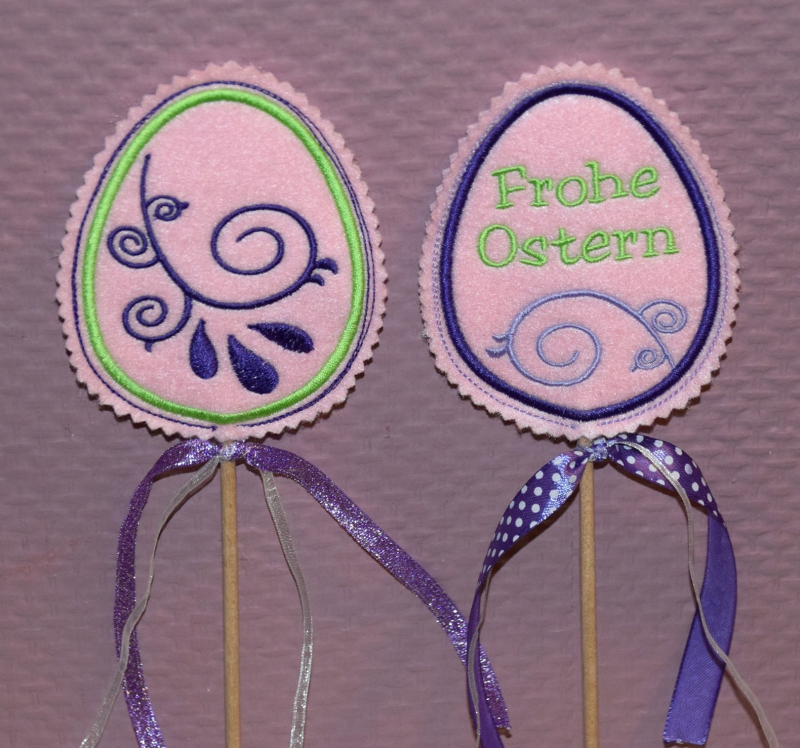 Stickdatei Set ITH Osterei Anhänger & Stabdesigns, Foto mit 2 Designstäben in Ostereiform. Bestickung auf rosa Untergrund: Schnörkelverzierung, 1 mal zudem Schriftzug "Frohe Ostern". Zierbändchen an Stäben zur Deko.