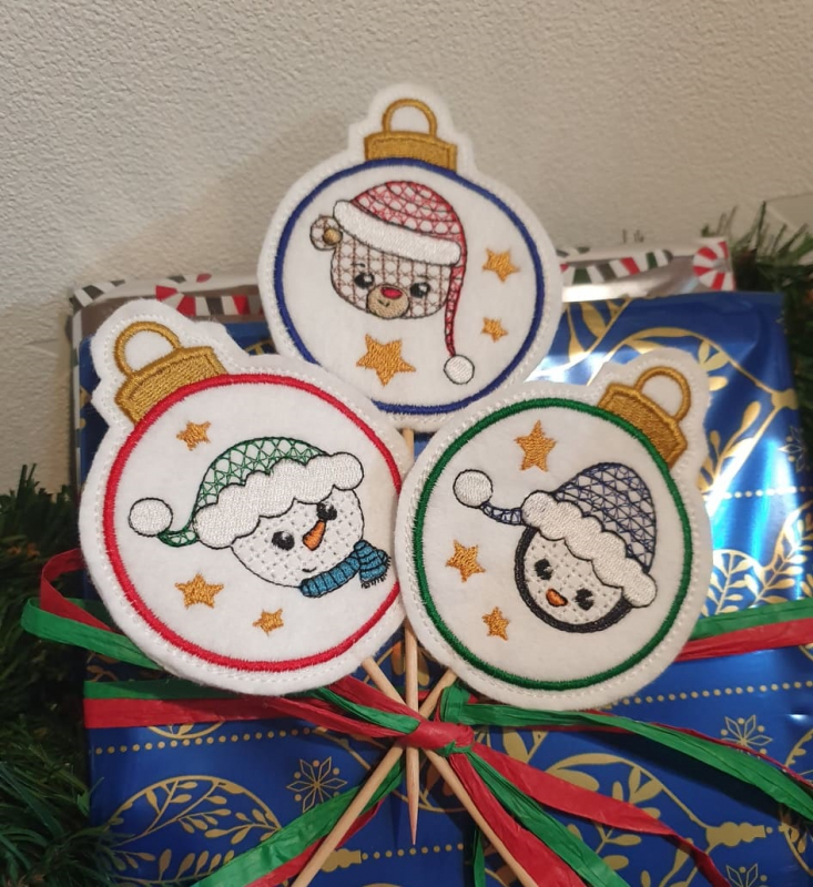 Stickdatei Set ITH Stecker Christmas Faces für Schaschlikspieße, Foto mit 3 Dekostäben, in die Schleife einer Weihnachtsgeschenkbox gesteckt. Motive: Weihnachtsbaumkugeln; Bestickung mit Schneemann-, bzw. Teddybärenkopf mit Mütze, Sterne.