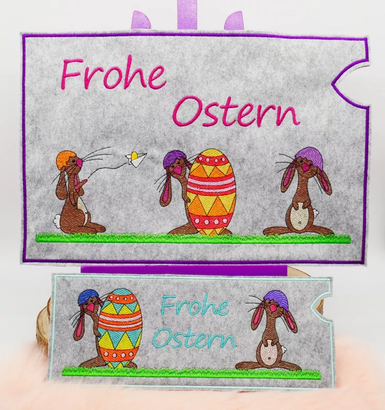 Stickdatei Set Hase Jasper ITH Geschenk- und Schokoladenhüllen inkl. Stabdeko, Foto zeigt 2 hellgraue querformatige Filzhüllen, bunt bestickt. Beide mit Schriftzug "Frohe Ostern". Größere mit unterschiedlichen 3 Hasen und Osterei, kleinere mit unterschied