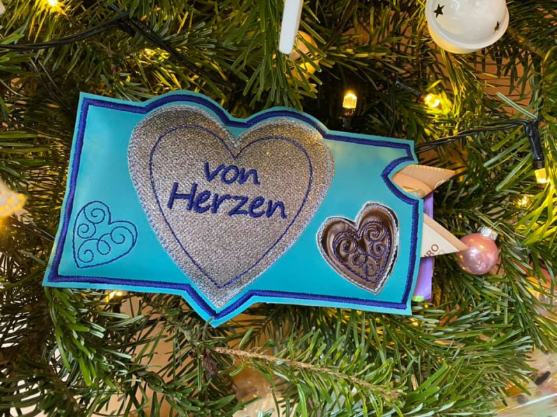 Stickdatei Set ITH Gutschein- und Schokohüllen Danke und Geburtstag, Foto zeigt Hülle vor Weihnachtsbaumhintergrund, gefaltete Geldscheine und lila Papier ragen aus ihr heraus. Blaue Querformat-Hülle mit 3 Herzen als Applikation in bronze und grau und als