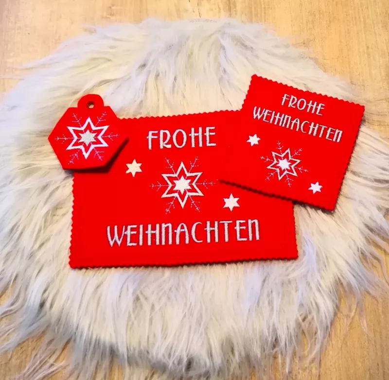 Stickdatei Set ITH Weihnachtskarten, Geschenkanhänger und Schriftzüge. Foto präsentiert auf hellem Fell 2 Weihnachtskarten und 1 geösten Geschenkanhänger in rot. Anhänger mit Eisstern-Bestickung; Karten mit Sternenbestickung und Schriftzug "Frohe Weihnach