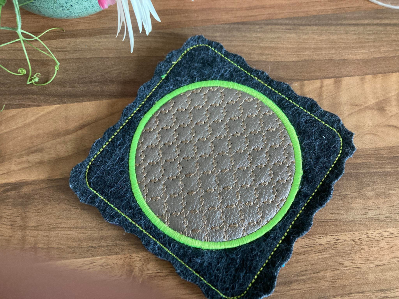 Stickdatei Set ITH Mug Rug 10. Der Ausschnitt zeigt auf einem Holzuntergrund den Mug Rug als quadratischen dunkelgraublauen wellig geschnittenen Filz mit hellgrün abgestepptem Rand. Mittig ein wellig-blattartiges Ornament in grau, das mit dem hellgrünen Stickdatei Set ITH Mug Rug 10. Der Ausschnitt zeigt auf einem Holzuntergrund den Mug Rug als quadratischen dunkelgraublauen wellig geschnittenen Filz mit hellgrün abgestepptem Rand. Mittig ein wellig-blattartiges Ornament in grau, das mit dem hellgrünen