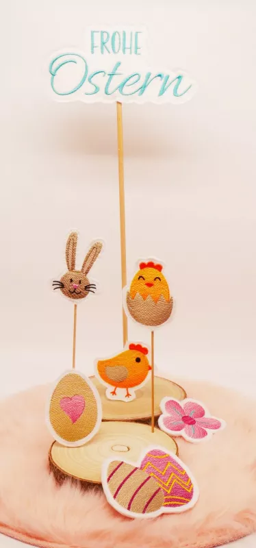 Stickdatei Set Oster-Stabdesigns, Foto mit 7 Designs auf 2 Holzscheiben, die auf orangem Fell liegen. Hasenkopf; laufendes Küken; Küken in Eierschale; Blüte; Ei mit Herz darin; 2 Ostereier; Schriftzug "Frohe Ostern".