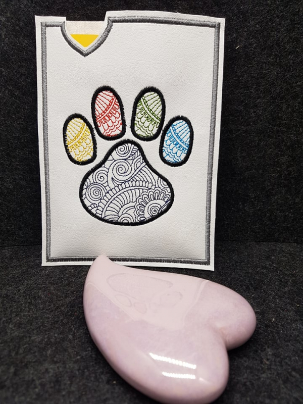 Stickdatei Set ITH TIERPASS-Hüllen 13 Designs (13x18), Foto mit Hülle vor dunkelgrauem Hintergrund, davor liegt ein herzförmiger rosa Stein. Helle Hochkanthülle mit großem Pfotenabdruck, der innen in diversen Farben eine filigrane Musterbestickung aufweis Stickdatei Set ITH TIERPASS-Hüllen 13 Designs (13x18), Foto mit Hülle vor dunkelgrauem Hintergrund, davor liegt ein herzförmiger rosa Stein. Helle Hochkanthülle mit großem Pfotenabdruck, der innen in diversen Farben eine filigrane Musterbestickung aufweis