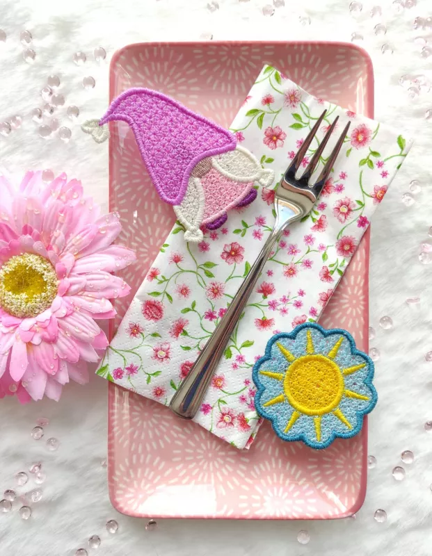 Stickdatei Set FSL Gnome rund um das Jahr + ITH Anhänger, Foto zeigt 2 ausgestickte Einzelmotive, in netzartiger Optik gefertigt. Gnom in pink/rosa/lila/weiß/braun; gelbe Sonne auf wellig-rundem Untergrund in mittelblau.Stickdatei Set FSL Gnome rund um
