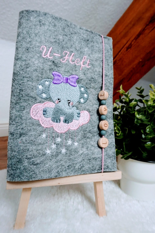 Stickdatei Set Elefanten (Vollstick) - ideal für Geschenke zur Geburt, Foto mit Ausfertigung, präsentiert auf Staffelei. Auf graumelierter Filzhülle Stickerei mit Elefant auf Wolke mit Sternchen, darüber Schriftzug "U-Heft". Als Hüllenverschluss Gummischn