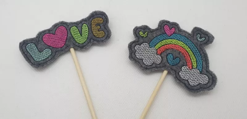 Stickdatei Set Valentinsmöpse ITH Stabdesigns für Stabdeko, Foto mit zwei Designs auf jeweils anthrazitfarbenem Filzuntergrund. Einmal bunter Schriftzug "LOVE" mit rotem Herz als "O", einmal bunter Regenbogen, von Herzen umgeben und mit je einer Wolke an 