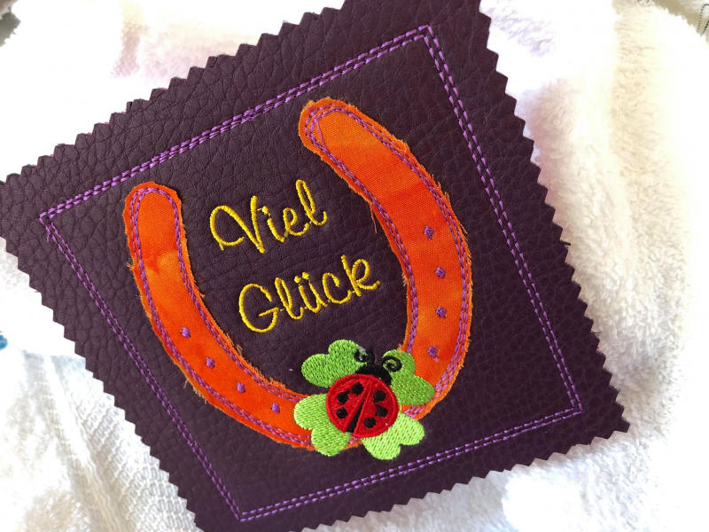 Stickdatei Set ITH Mini-Grußkarten inkl. Abwandlung zur Geschenktasche, Foto mit quadratischer Ausarbeitung, auf hellem Untergrund liegend. Auberginefarbenes Kunstleder mit Bestickung: Schriftzug "Viel Glück" in einem Hufeisen, das mit Marienkäfer auf Glü Stickdatei Set ITH Mini-Grußkarten inkl. Abwandlung zur Geschenktasche, Foto mit quadratischer Ausarbeitung, auf hellem Untergrund liegend. Auberginefarbenes Kunstleder mit Bestickung: Schriftzug "Viel Glück" in einem Hufeisen, das mit Marienkäfer auf Glü