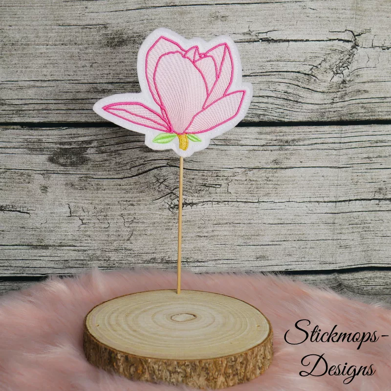Freebie Stickdatei ITH Stabdatei Magnolie, Foto einer Ausfertigung, die mit dem Stab in einer Holzscheibe steckt. Stoffuntergrund aus hellem Filz in Blütensilhouette. Blütenzeichnung in rosa-rot mit filigraner rosa Ausstickung. Unterhalb der Blüte Stilans