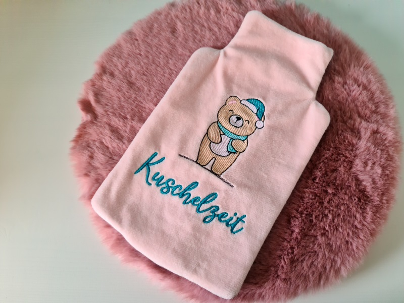 Stickdatei XXL Set ITH Wärmflaschenhüllen mit 24 Designs, Foto einer Hülle aus apricotfarbenem Flauschstoff. Darauf ein Teddybär mit Mütze und Schal, unten Schriftzug "Kuschelzeit".n und Sternen.