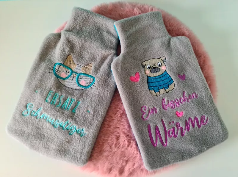 Stickdatei XXL Set ITH Wärmflaschenhüllen mit 24 Designs, Foto zeigt 2 Hüllen aus grau-braunem Flausch-Stoff. Einer mit  Katzenkopf, darunter Schriftzug "Ersatz-Schmusetiger". Der andere mit Mops und Herzchen, Schriftzug "Ein bisschen Wärme".
