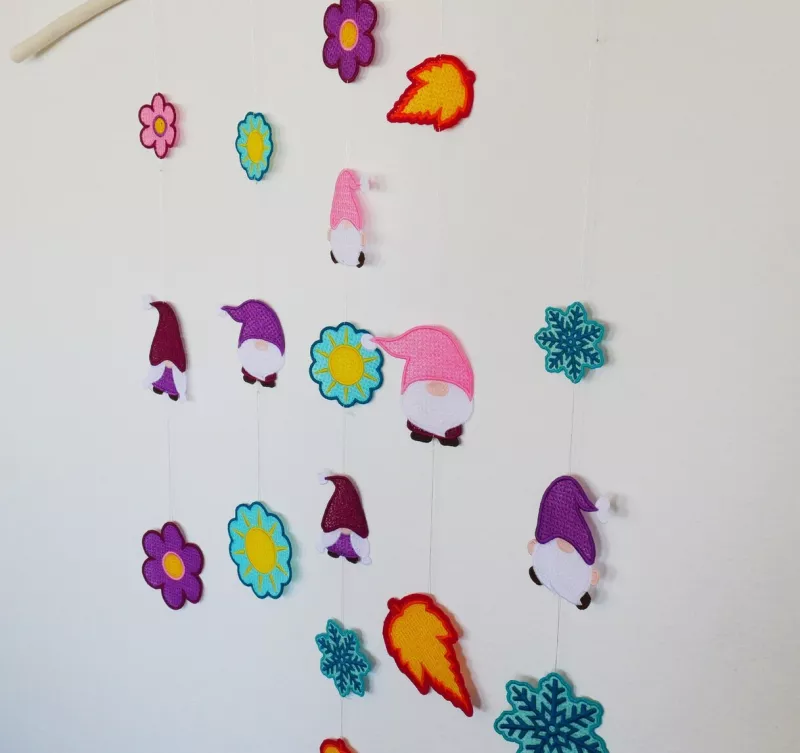 Stickdatei Set FSL Gnome rund um das Jahr + ITH Anhänger, Foto zeigt 18 ausgefertigte bunte Anhänger. 3 mal stilisierte Blüten, 3 mal Sonne, 3 mal Schneekristall, 6 mal Gnom3, 3 mal Laubblätter. Teilweise Varianzen in Farbkombinationen und Größe.