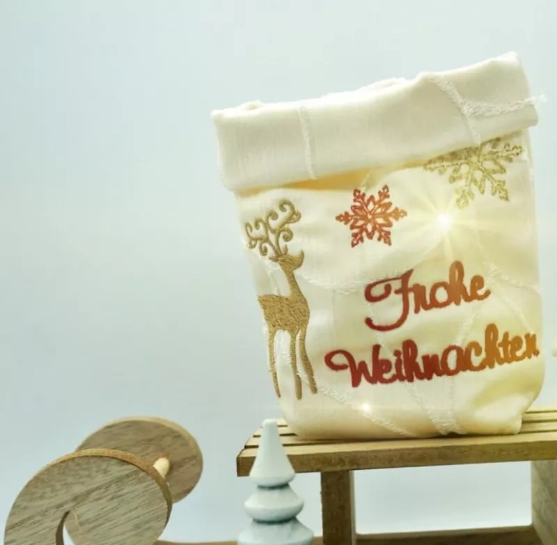 Stickdatei Set 6 winterliche Lichtbeutel (für den 13x18 Rahmen), Foto mit Lichtbeutel aus hellem Stoff mit umgeschlagenem Rand. Bestickung in gold und rot: Hirsch, Eiskristalle, Schriftzug "Frohe Weihnachten".
