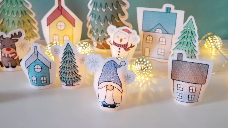 Stickdatei Set ITH LED Cover Winterdorf mit Gnomen, mit Bäumen und Schneemann. Man sieht eine Lichterkette als Dekoelement.