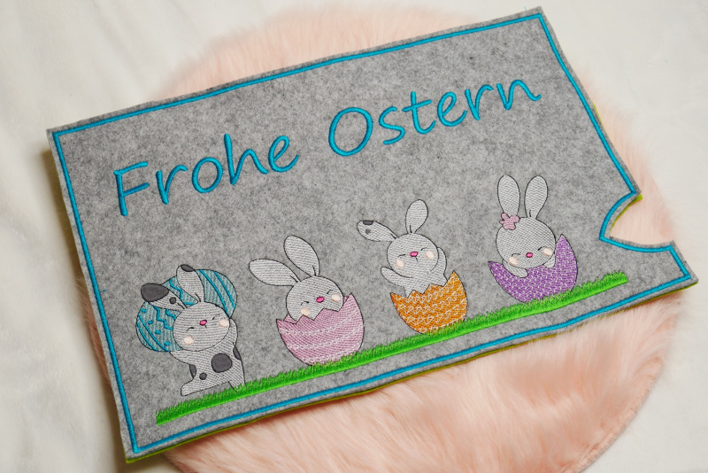 Stickdatei Set Osterhüllen ITH Geschenk- und Schokoladenhüllen + Einzelmotive. Foto, das eine Ausfertigung auf apricotfarbenem Fell liegend zeigt. Querformatige Hülle aus grauem Filz mit Schriftzug "Frohe Ostern", darunter 4 verschiedene Osterhasenmotive 