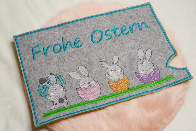 Stickdatei Set Osterhüllen ITH Geschenk- und Schokoladenhüllen + Einzelmotive. Foto, das eine Ausfertigung auf apricotfarbenem Fell liegend zeigt. Querformatige Hülle aus grauem Filz mit Schriftzug "Frohe Ostern", darunter 4 verschiedene Osterhasenmotive 